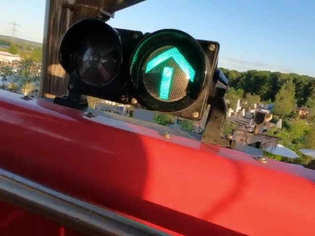 A green traffic arrow is illuminated against a clear blue sky from a red vehicle window frame, offering a high view of a suburban landscape. The bright signal stands out in the foreground over green trees and rooftops.