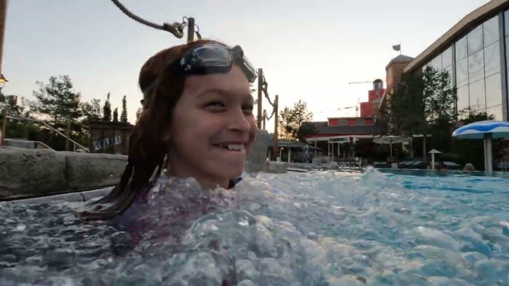 A person wearing goggles smiles while enjoying the water, with a modern building and lagoon in the background.