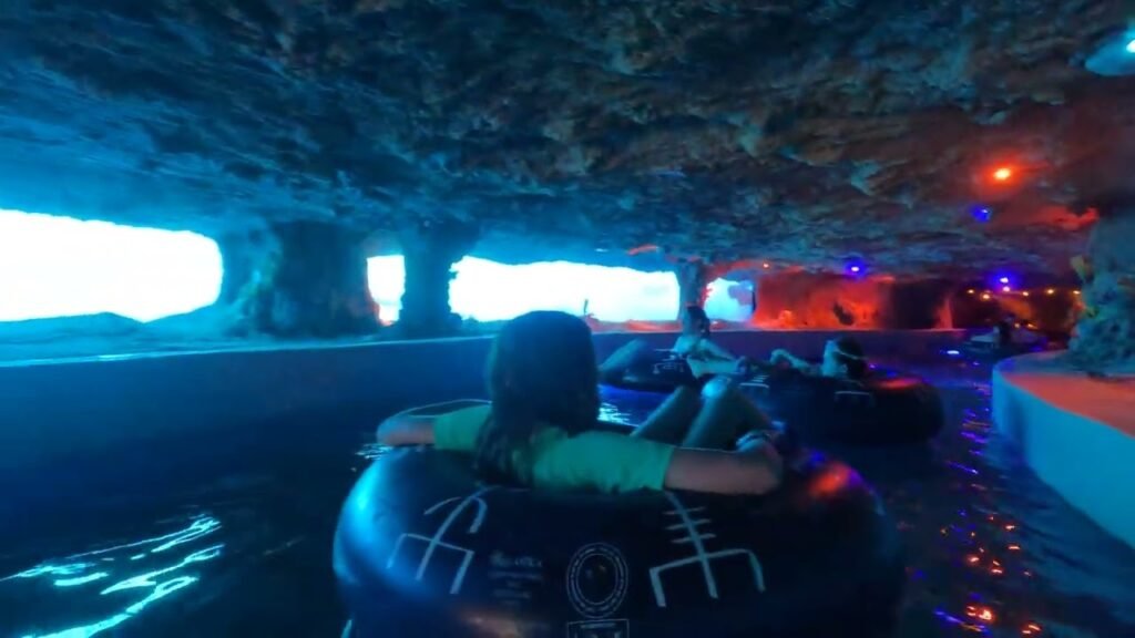 This image shows an indoor theme park ride called a 'Grotto Run' in a dark, cave-like environment with glowing lights and air tracks, where people float on inflatable tubes through a black lake.
