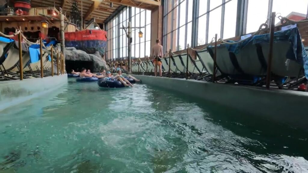 Guests relaxing on inner tubes drifting in an indoor river under a glass-roofed building with marine decor.