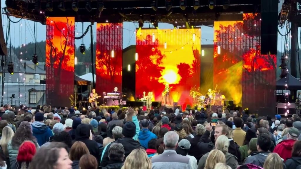 A large crowd enjoys a concert with colorful stage screens, set against a scenic backdrop.