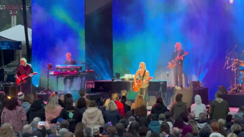A live band performs on an outdoor concert stage under vibrant blue and purple lighting.