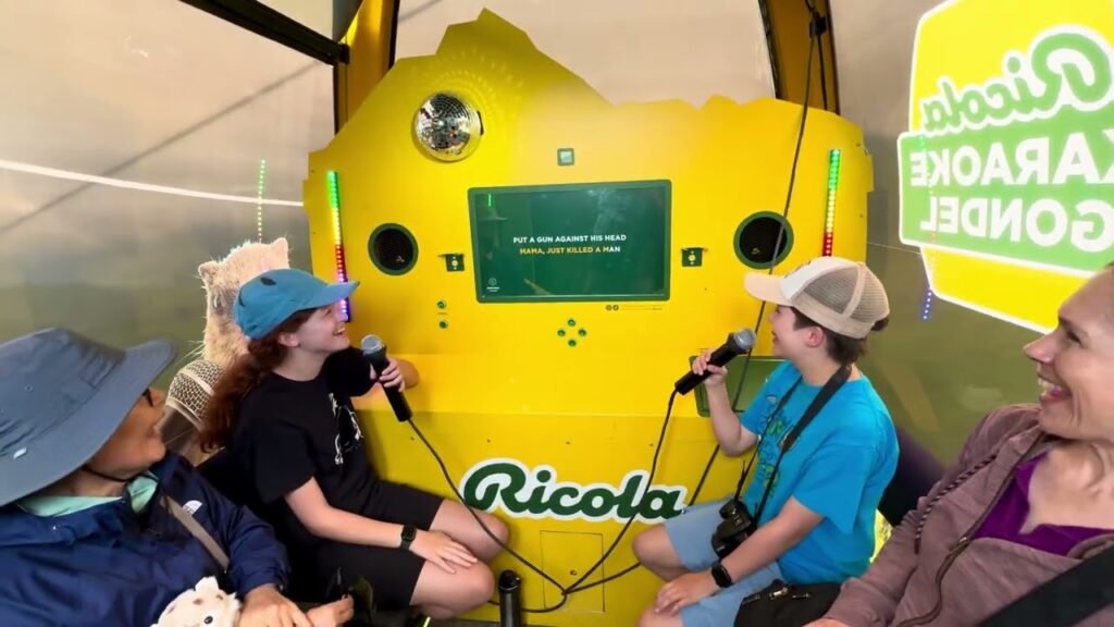 Passengers singing into microphones on the Ricola Gondel Cable Car in a bright yellow cabin featuring a 'Ricola' logo, during a scenic tourist excursion.
