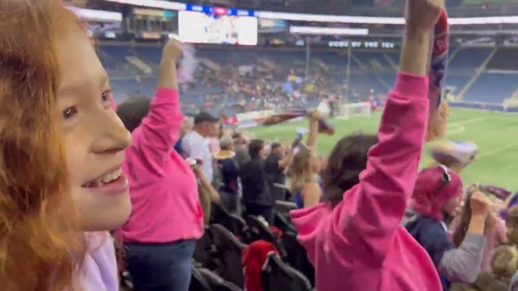 A person with red hair smiles excitedly in a baseball stadium crowd wearing a pink shirt, with other fans and the field visible in the background.