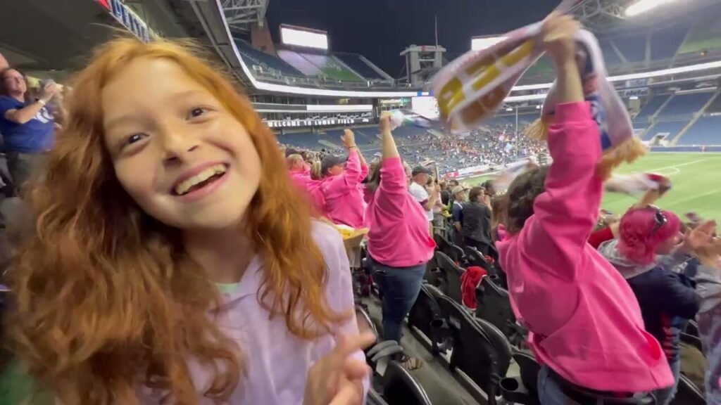 A girl smiles at the camera at a stadium while fans hold up a flag.