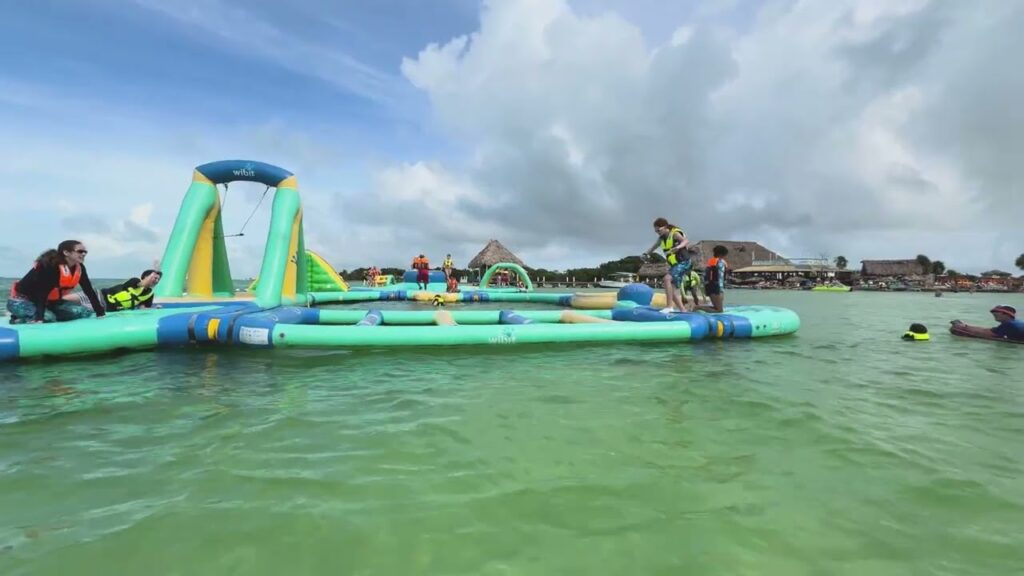 People playing on a colorful floating aqua park in clear shallow water with sandy shores and resort overwater bungalows in the background.