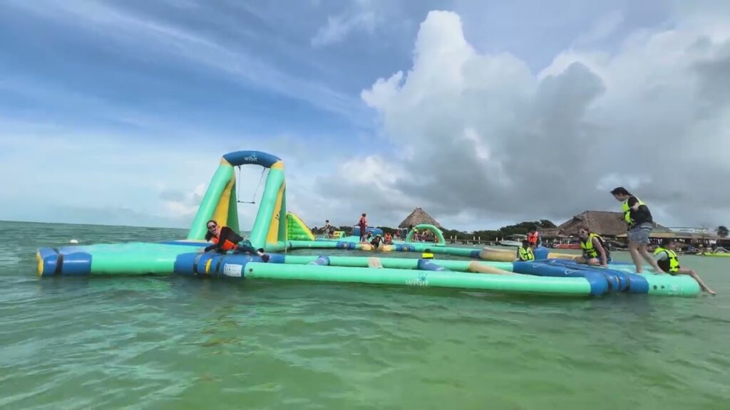 An inflatable water rally with a slide on calm green waters, surrounded by a tropical beach landscape and cloudy skies.