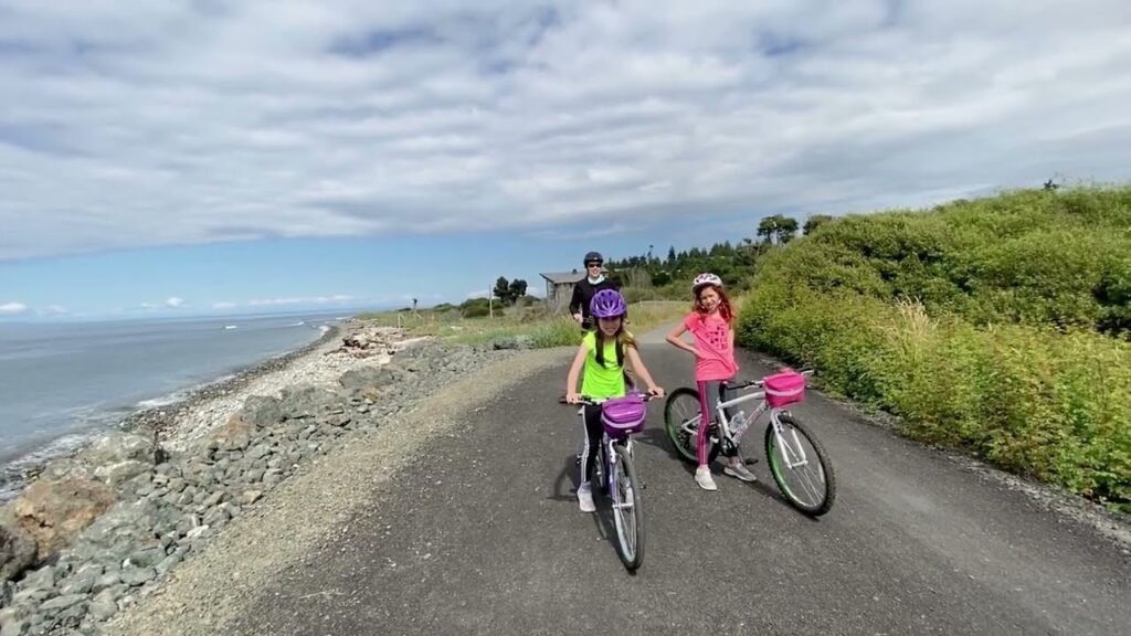 A family enjoying a scenic bicycle ride along a paved path in the Pacific Northwest, with lush greenery on one side and the ocean visible in the distance.