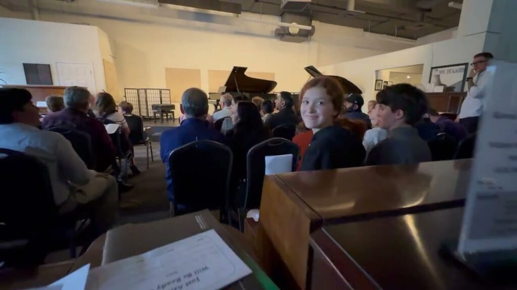 Audience members seated in a hall with a grand piano visible on stage during a community event.