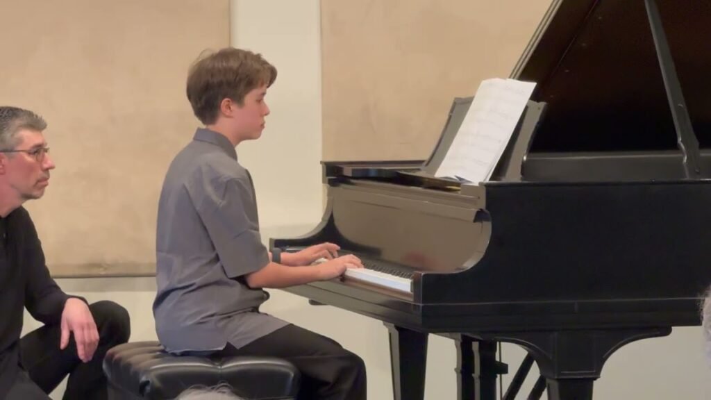 A young person playing a black grand piano in a recital setting, with another individual seated nearby.