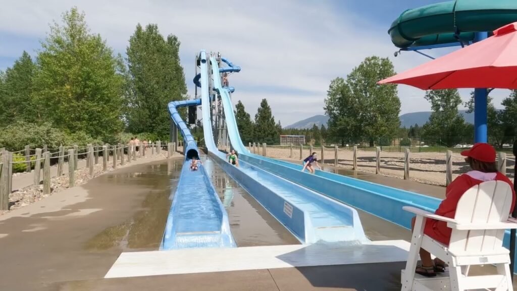 A view of a large blue water slide with a person in a red vest watching from a deck chair under an umbrella.