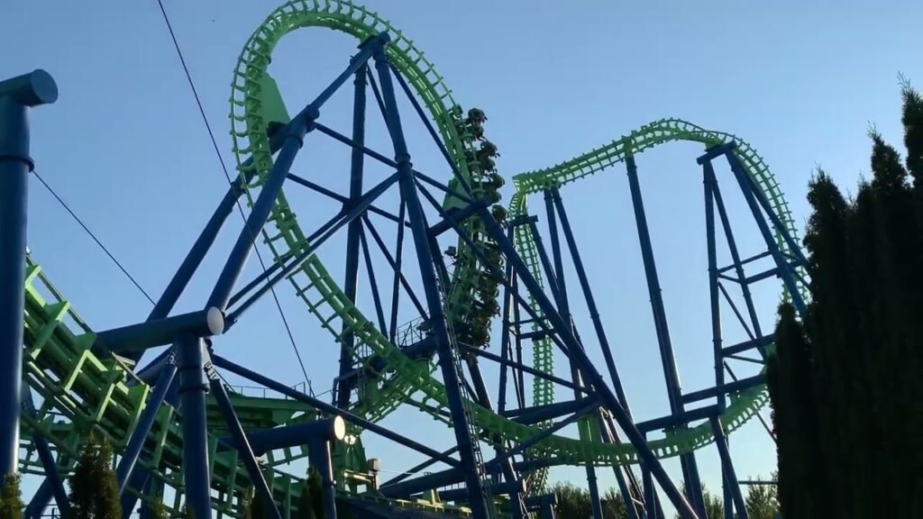A vibrant green and blue roller coaster with dramatic loops and twists set against a clear blue sky, framed by tall evergreen trees.