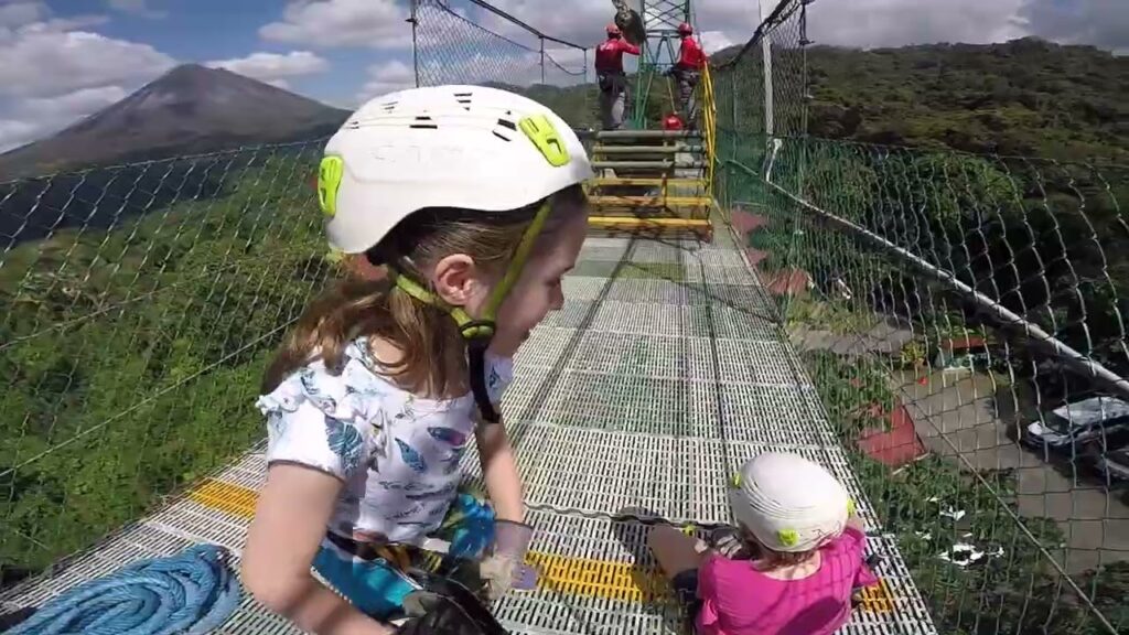 Children wearing helmets explore a metal mesh platform surrounded by safety netting and lush tropical vegetation.