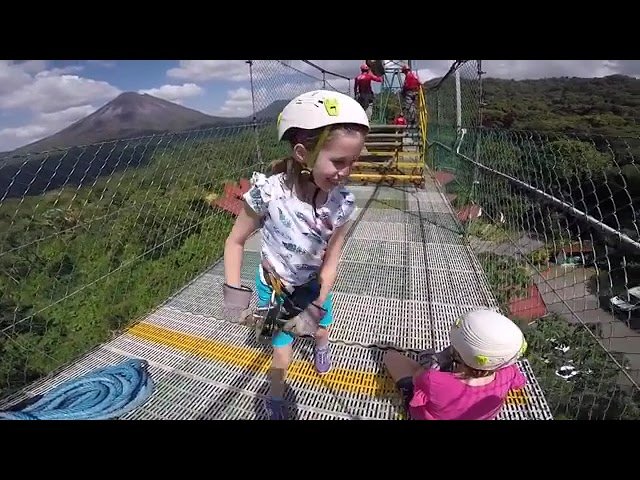 A person wearing a helmet and harness rides a zipline through lush green mountains. A mountain is visible in the background.
