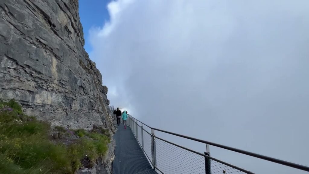 A high-altitude mountain trail winds along a steep rocky cliff face under a cloudy sky. Hikers walk along the narrow path equipped with safety railings.