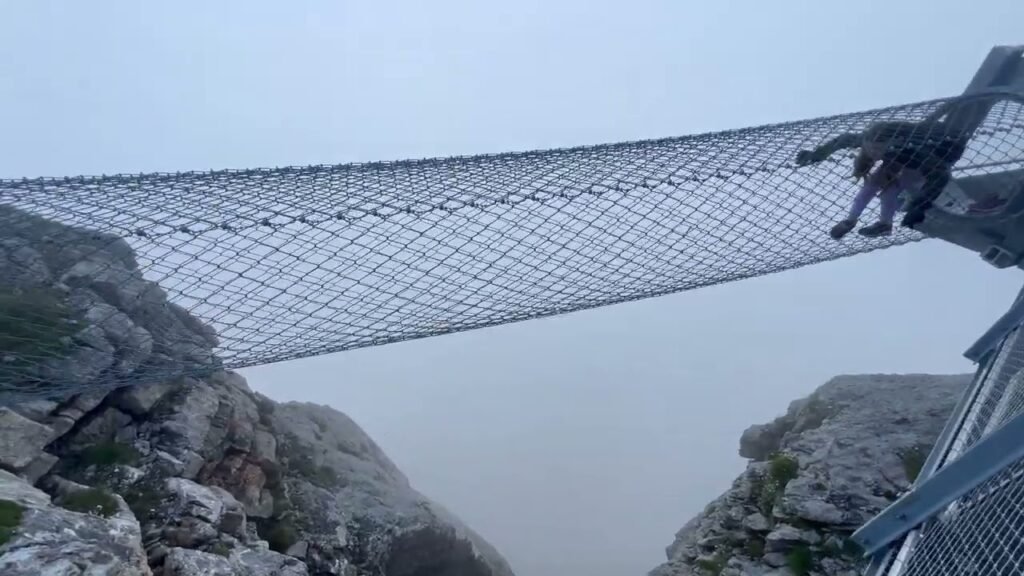 A cable bridge stretches across rocky cliffs under thick fog, capturing the thrill of an extreme European adventure.