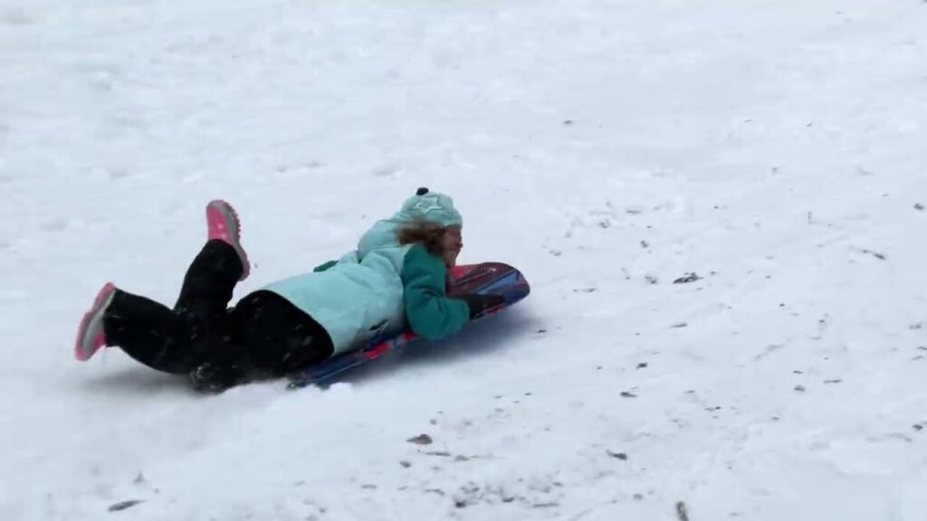 A person in a light jacket and pink shoes lays prone on a snowy slope. They are using a low-profile sled for a casual winter slide.