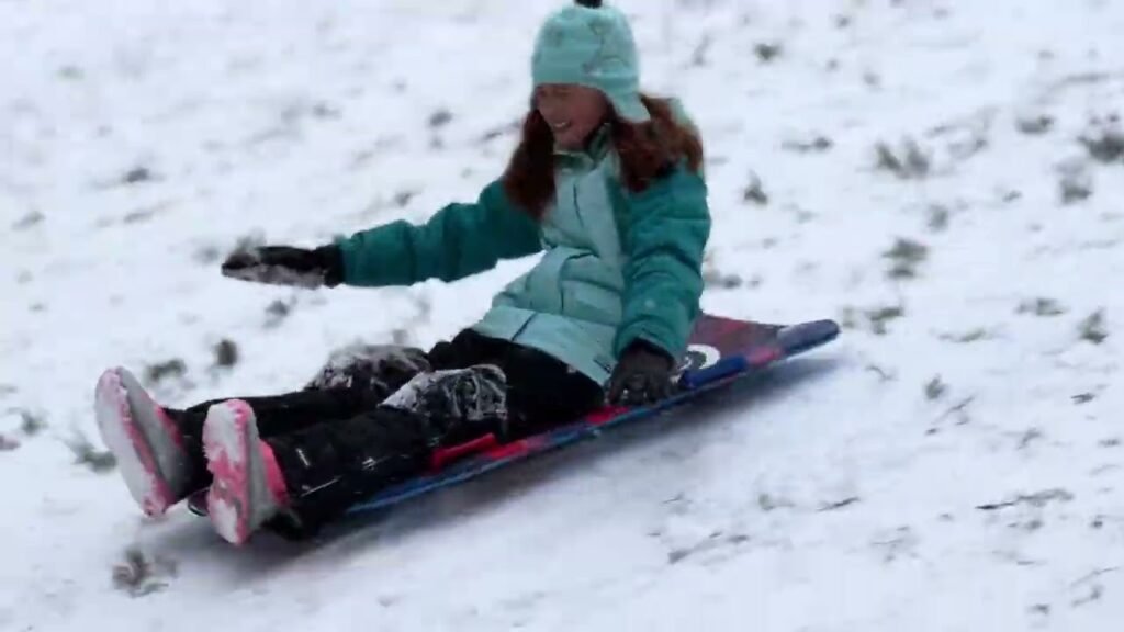 A child in a teal coat and beanie seated on a sled in the snow.