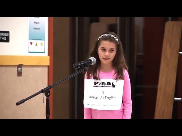 A child in a pink top speaks into a microphone at what appears to be a PTA event. The setting includes an indoor backdrop with a door, including the speaker's name badge visible.