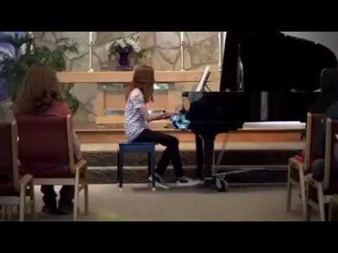A person plays a grand piano in a softly lit indoor space with an audience seated nearby.
