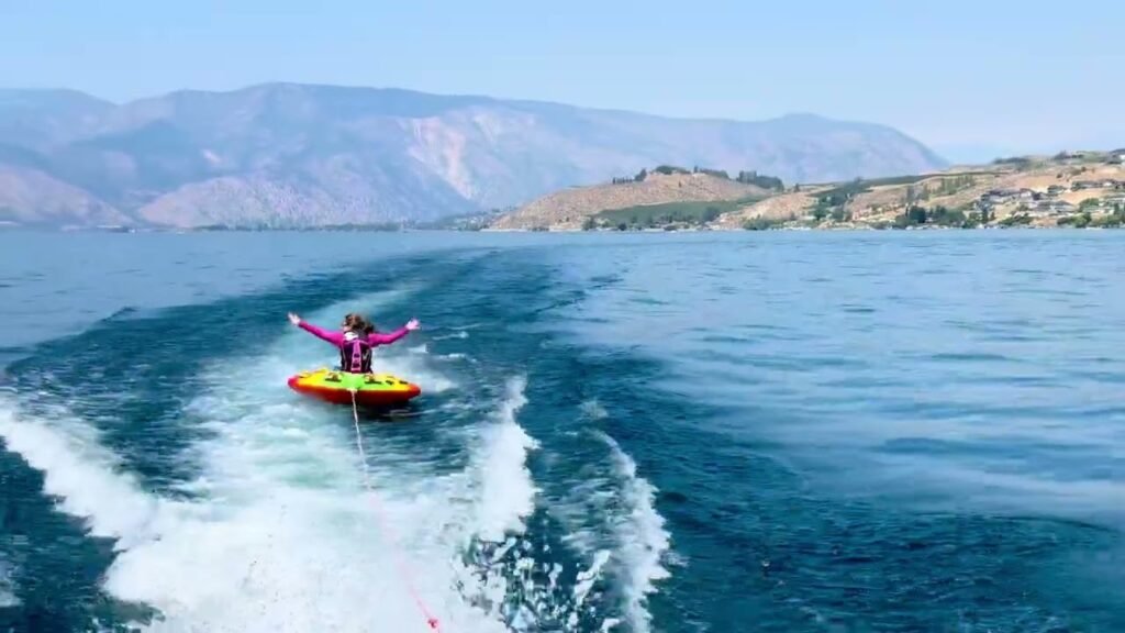 A person rides a red and yellow tube behind a boat on a deep blue lake with mountains in the background.