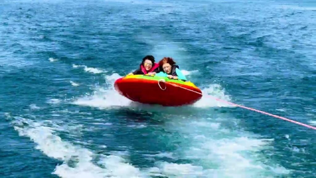 Two people enjoy an exciting ride in a bright red wake sled on open blue water. The sled is being towed across the ocean.