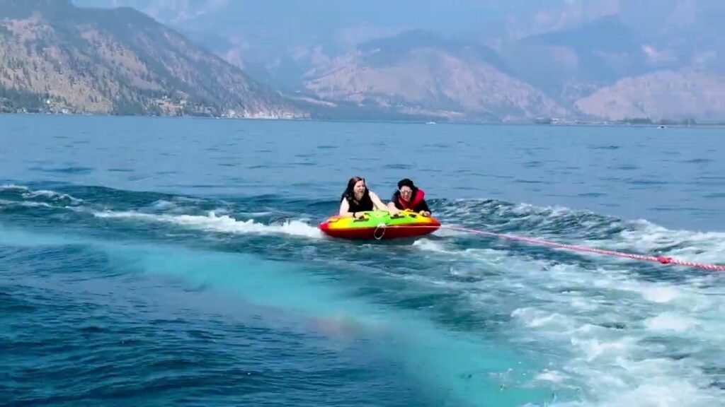 Two people in an inflatable tube are towed by a boat across Lake Votic, with mountains in the background.