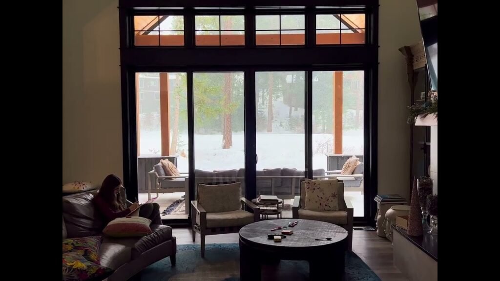 A cozy living room with large windows overlooking a snowy outdoor patio. The room features warm lighting and comfortable furniture.