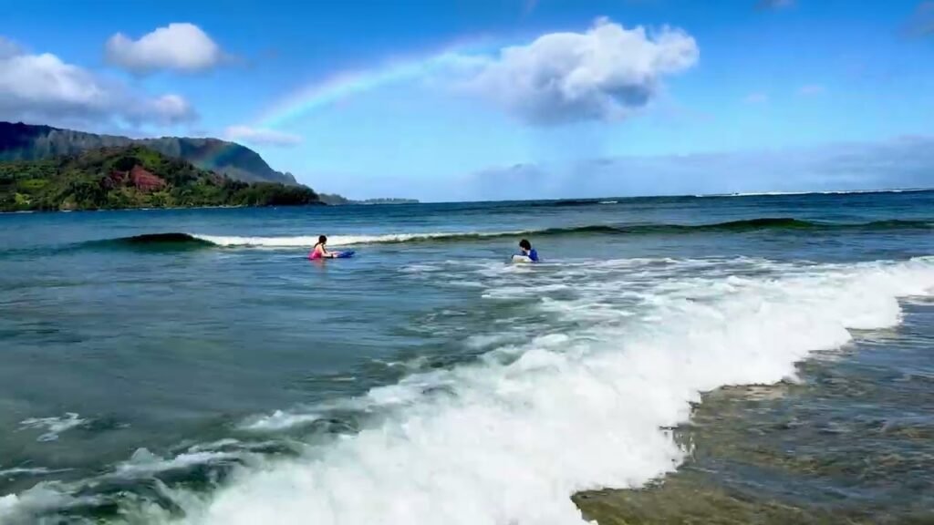 People surf on small curls breaking on a beach with green rolls in the background.