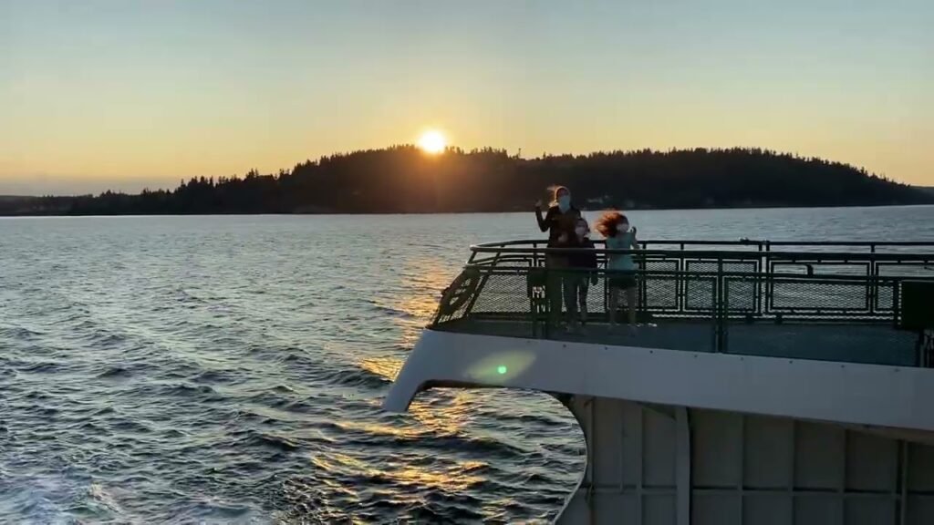 The sun sets behind a forested hill viewed from a cruise ship. The golden light reflects on the calm water surface.