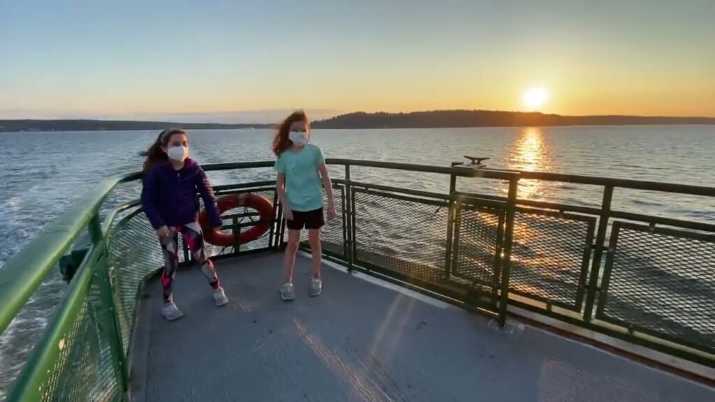 Two people in face masks standing on a boat deck at sunset in the Pacific Northwest. A calm ocean view with a low sun on the horizon.