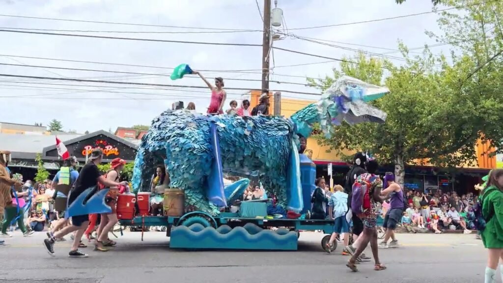 A vibrant blue and green dragon parade float rolls down a local street in Southeast Asia while participants perform alongside. Spectators watch from the sides as the float moves through the community under an overcast sky.