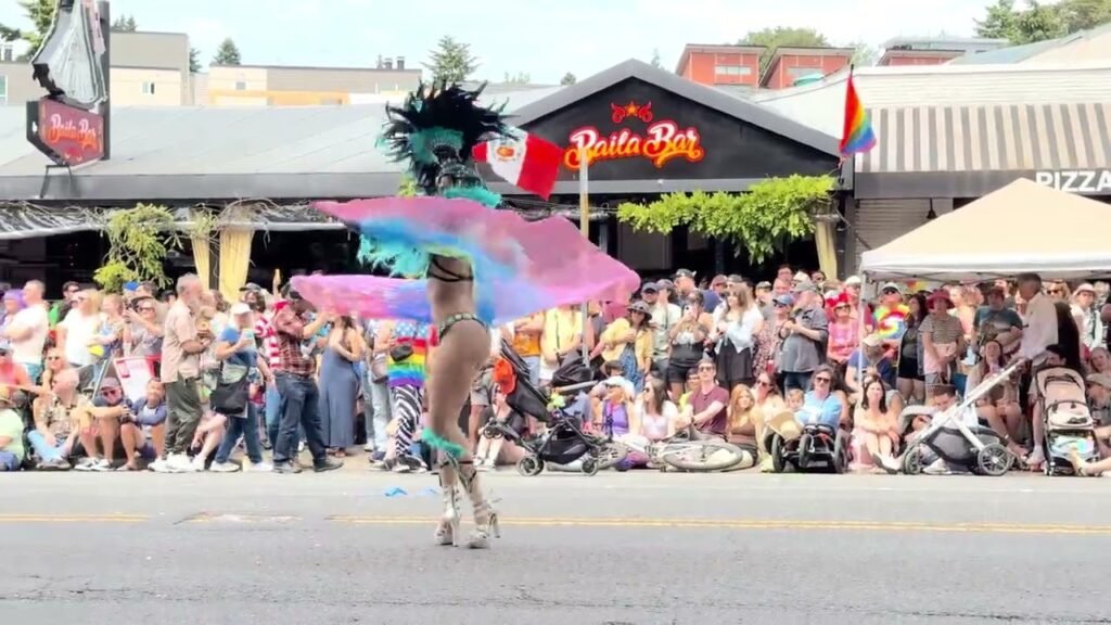 A participant holds a rainbow umbrella during a street parade. A crowd and a building with a rainbow flag are visible in the background.