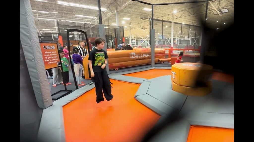 A child jumps on an orange trampoline in a busy indoor trampoline park surrounded by padded walls and other jumpers.