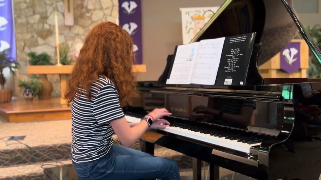 A woman with red hair plays the piano in a stone-walled room. The image highlights a grand piano with sheet music on the stand.