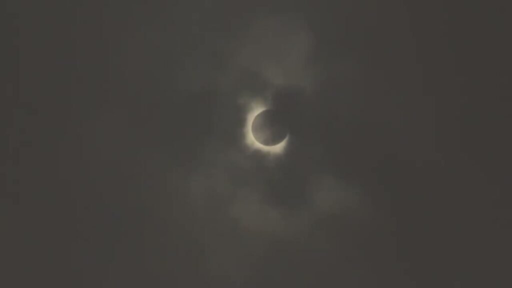 A crescent-shaped lunar eclipse captured through thin clouds. The moon glows brightly in a dark sky.