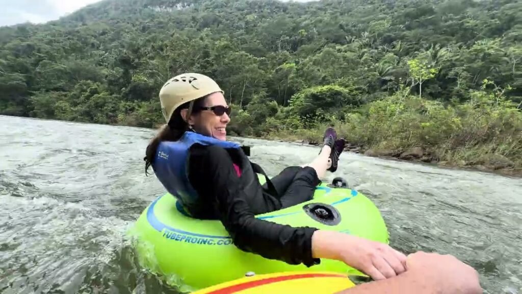 A person wearing a helmet and life vest relaxes on a bright green inner tube while floating down a river surrounded by lush forest.