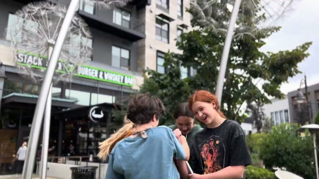 People chatting outside a city cafe with the words 'Stack' and 'Pittsburgh' visible in the background. A modern building and green trees surround the scene.