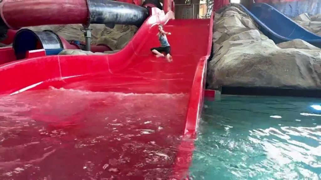 A bright red water slide at a waterpark leading into a turquoise pool. A person is sliding down the ramp next to a blue slide on artificial rock.