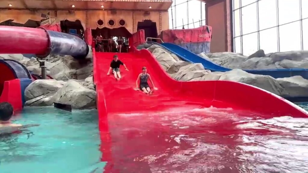 A wide red water slide at an indoor water park with people sliding down toward the pool, alongside a blue slide and rock features.
