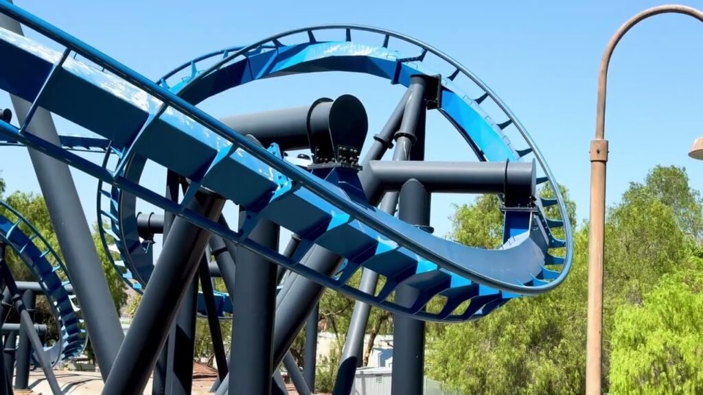 Blue coaster track curves dramatically against the sky in an amusement park. Bright blue rails and grey supports dominate the scene.