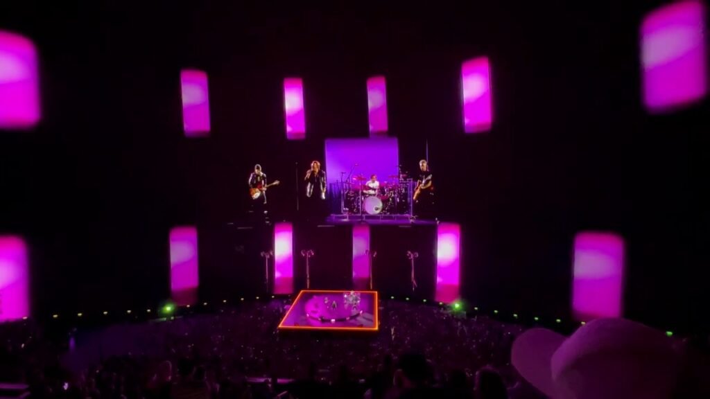 A band performs on a brightly lit stage with purple lighting and a large crowd in attendance at a live concert.