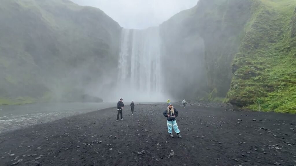 Tourists standing on dark volcanic gravel before the historic Skógafoss waterfall in Iceland. Lush green cliffs frame a torrent of water pouring into a mist-covered base.