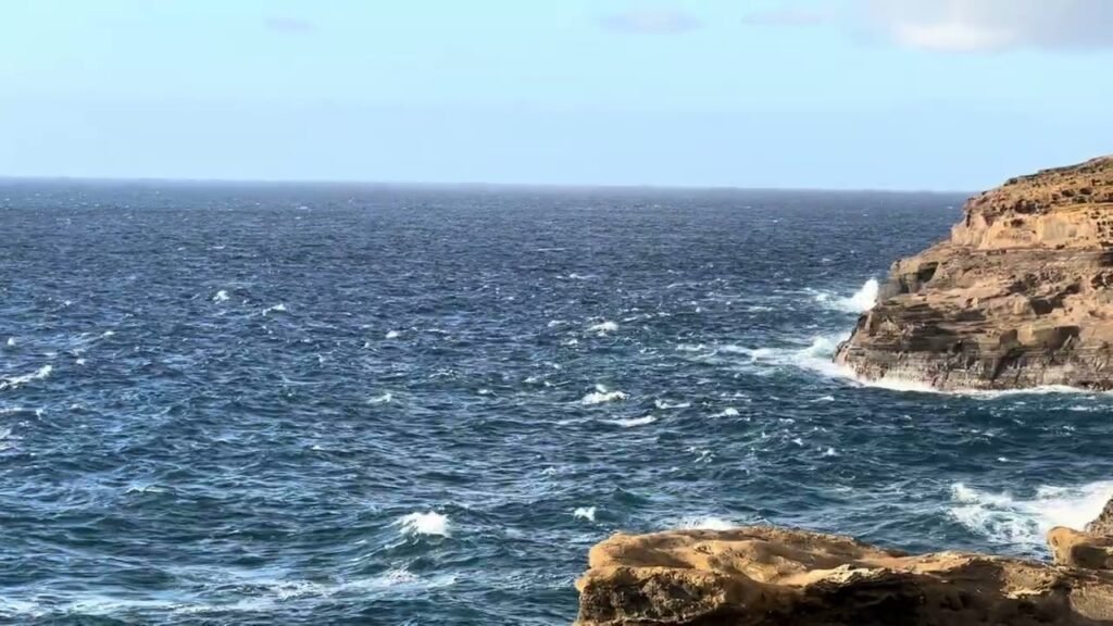 Waves crashing against rugged coastal cliffs with a vast deep blue ocean stretching to the horizon under a clear sky.