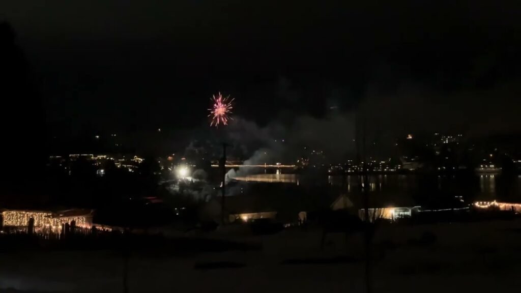 Red fireworks burst in the night sky above a waterfront city with illuminated buildings reflected in the dark water.