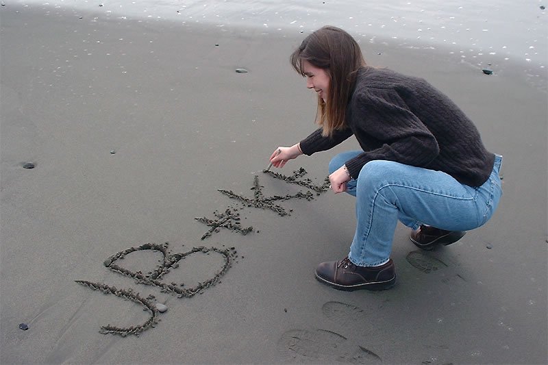 A person is writing the initials JB + K in the wet sand on the beach. The individual is wearing jeans and a black sweater.