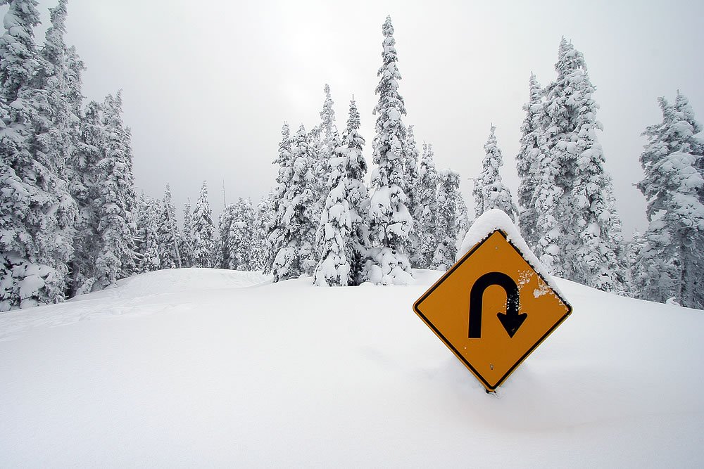 A yellow road sign with a hairpin curve arrow stands in deep white snow. Snow-covered evergreen trees loom in the background.