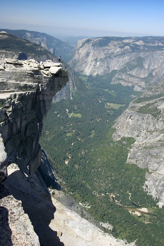 A steep cliffside view overlooking a lush green valley and distant mountains under clear blue skies. A lone figure stands near the rocky edge, emphasizing the vastness of the landscape below.