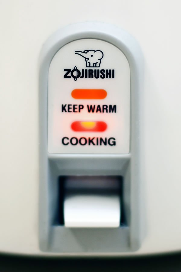 Zojirushi rice cooker with red LED lights indicating 'Keep Warm' and 'Cooking' statuses, featuring a digital display and user interface.